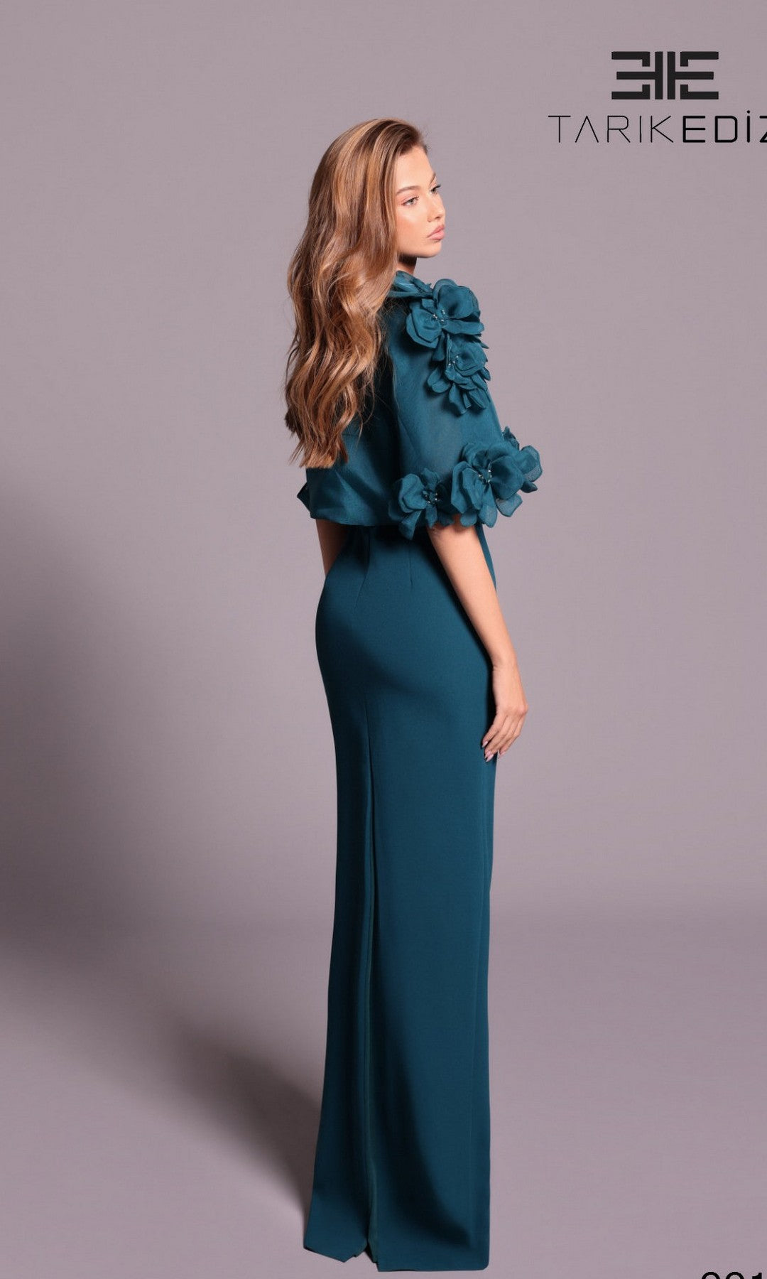 A woman in a teal Tarik Ediz 99134 long formal dress with 3D floral appliqué on the sleeves stands sideways against a plain background.